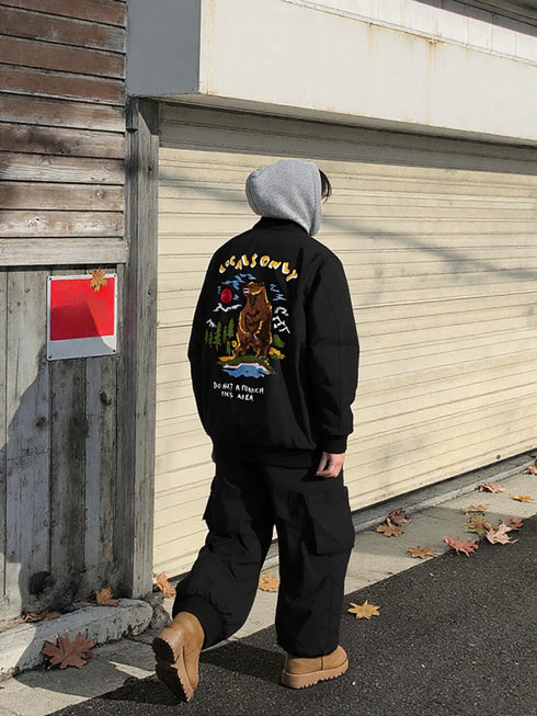 Thick embroidered baseball jacket in black with bear embroidery on back, worn with a hoodie and cargo pants for winter streetwear.