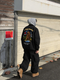 Thick embroidered baseball jacket in black with bear embroidery on back, worn with a hoodie and cargo pants for winter streetwear.