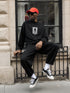 Man wearing black Japanese streetwear sweatshirt with graphic print, black pants, red cap, and sunglasses sitting on street railing