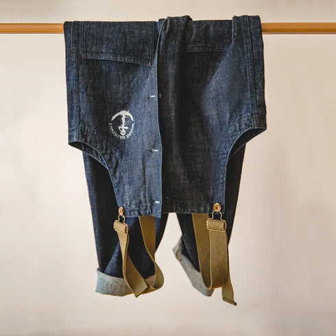 Blue jeans with suspenders hanging on a wooden rod against a beige background