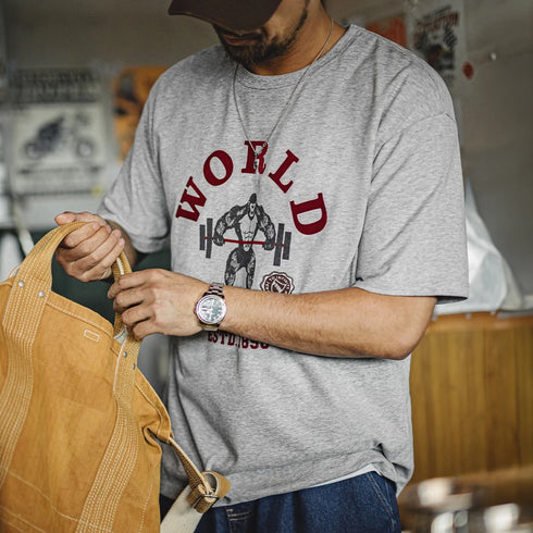 Person wearing a gray t-shirt with a graphic design, holding a tan jacket indoors.