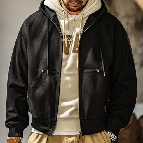 Person wearing a black jacket over a beige hoodie with a logo, standing against a neutral background.