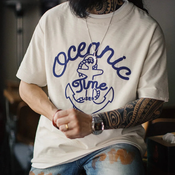 Person wearing a white t-shirt with 'Oceanic Time' design and tattoos on arms.