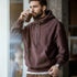 Man wearing a brown hoodie drinking from a glass indoors.