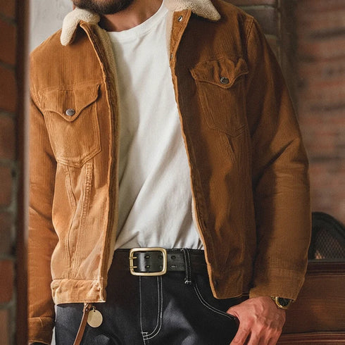 Person wearing a brown corduroy jacket with a white shirt and black pants against a brick wall.