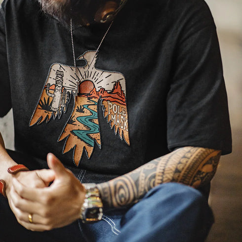 Person wearing a black t-shirt with a colorful eagle design and text, sitting with tattooed arms.