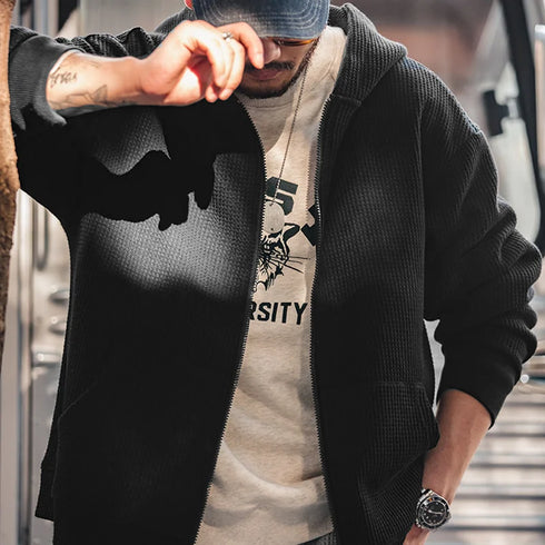 Person wearing a black cardigan over a beige hoodie with a logo, standing outdoors.