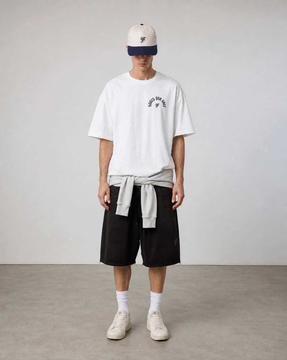 A model stands in a studio wearing a white t-shirt with a black graphic, black shorts, white socks, and white sneakers, with a light gray tie-worn shirt and a cream cap.