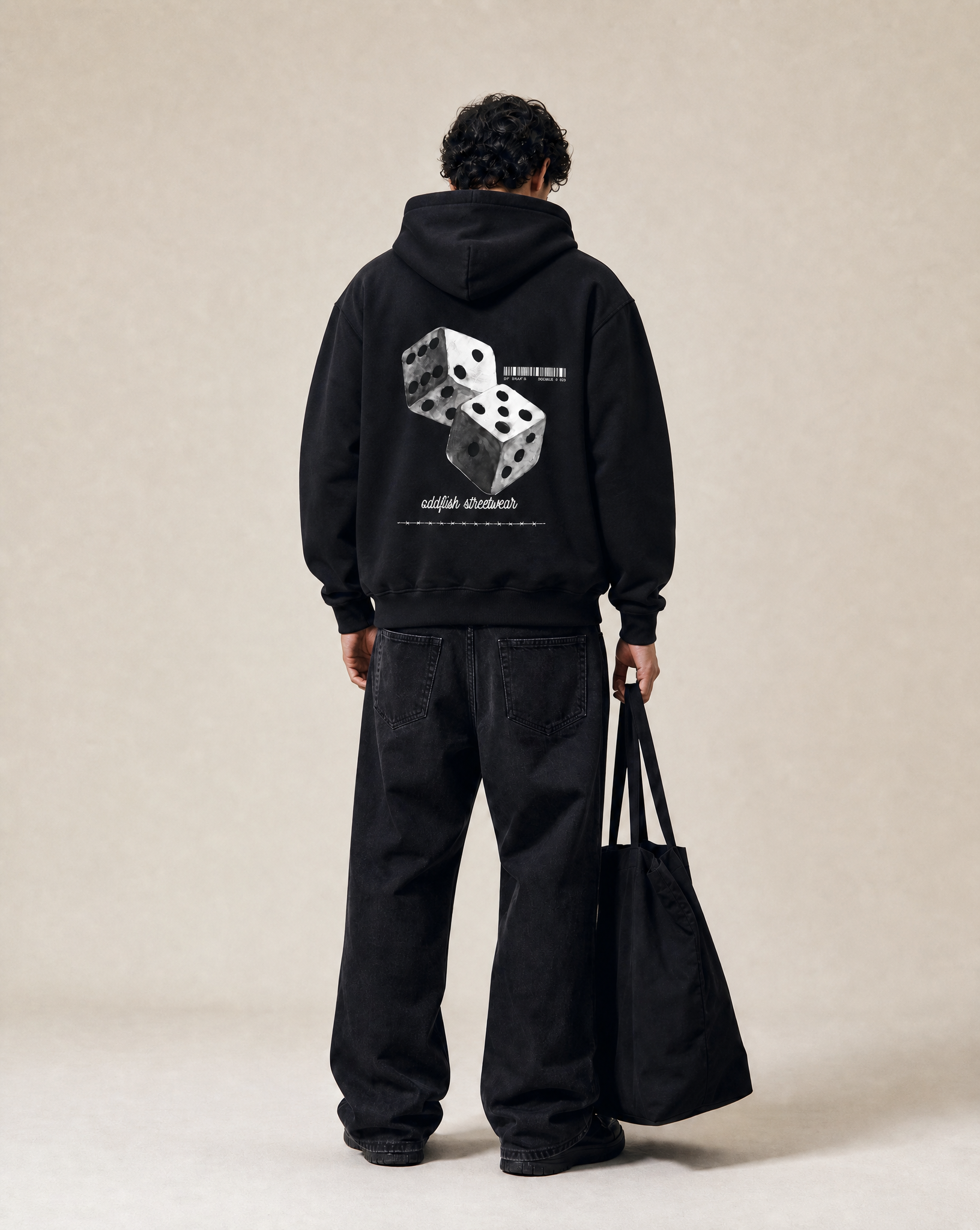 A model viewed from behind wearing a black hoodie with a large white graphic of two dice on the back, paired with black jeans and holding a black tote bag against a neutral beige background.