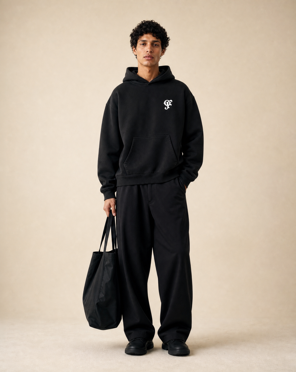 A man modeling a black hoodie and matching black sweatpants against a warm beige studio background, holding a black tote bag.