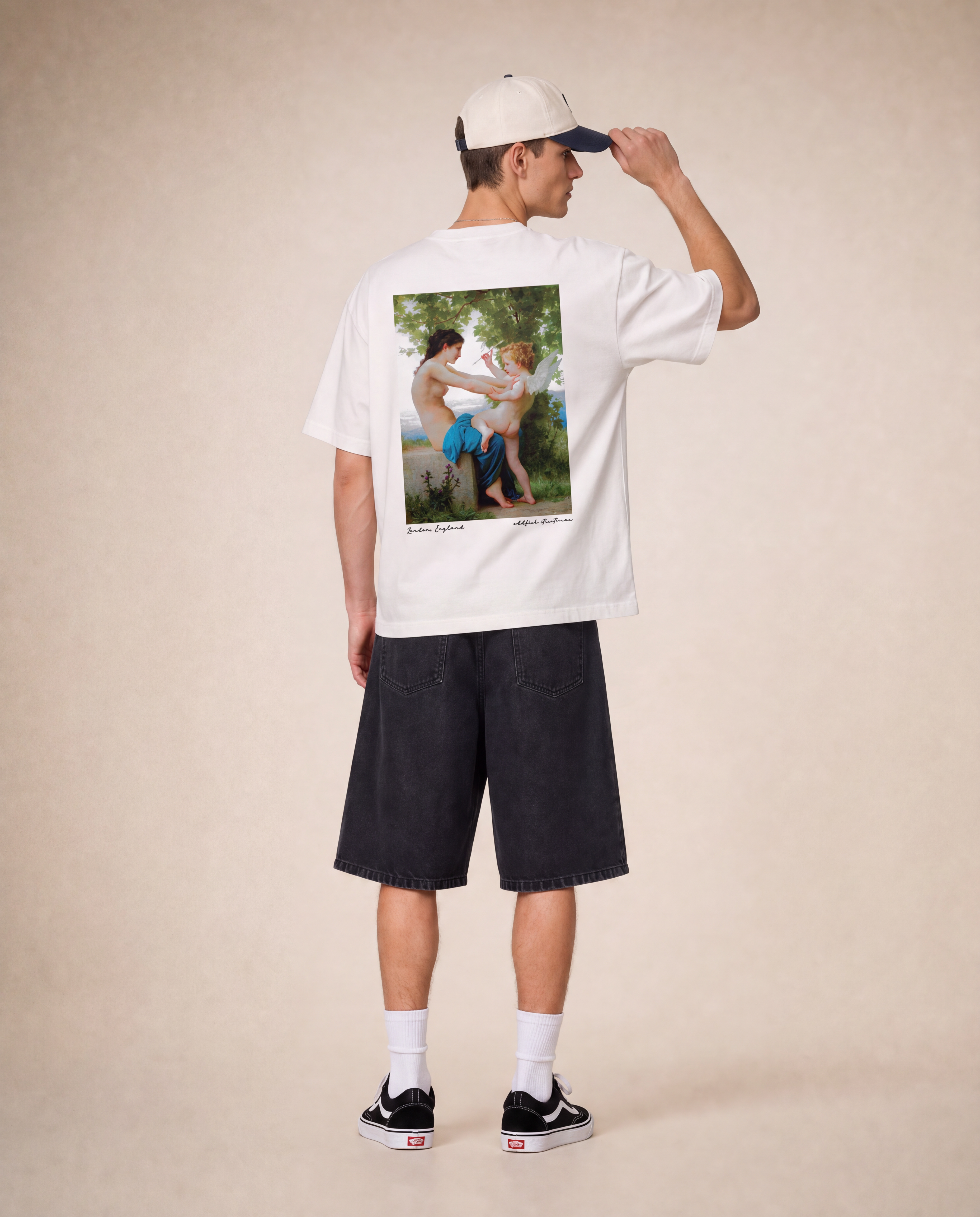 A model shown from the back wearing a white t-shirt with a large vintage-style photographic print on the back, paired with dark denim shorts, a cream baseball cap, white socks, and black sneakers, set against a warm beige studio backdrop.