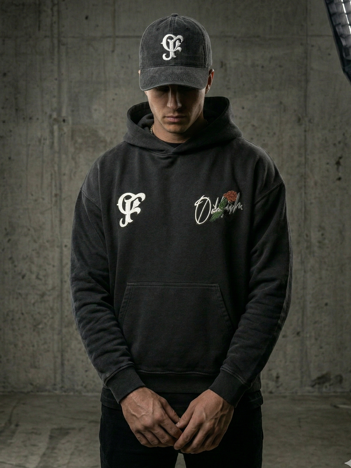 Person wearing a hoodie with logos and a cap in a concrete setting black gray blue