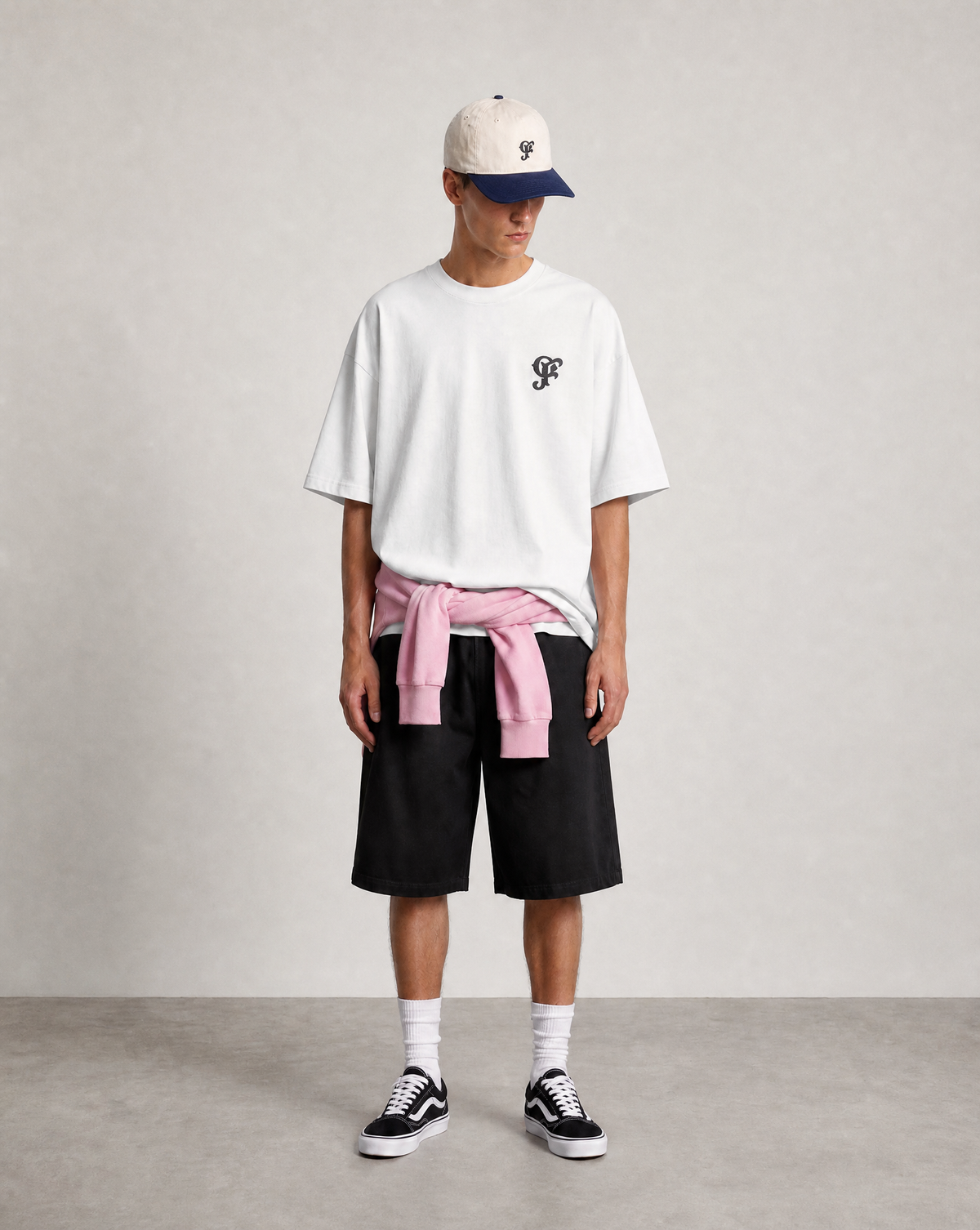 A model stands against a neutral light gray studio background wearing an oversized white t-shirt with a small black embroidered logo on the left chest, black shorts, a pink shirt tied around the waist, white socks, black and white sneakers, and a cream baseball cap with a navy blue brim.