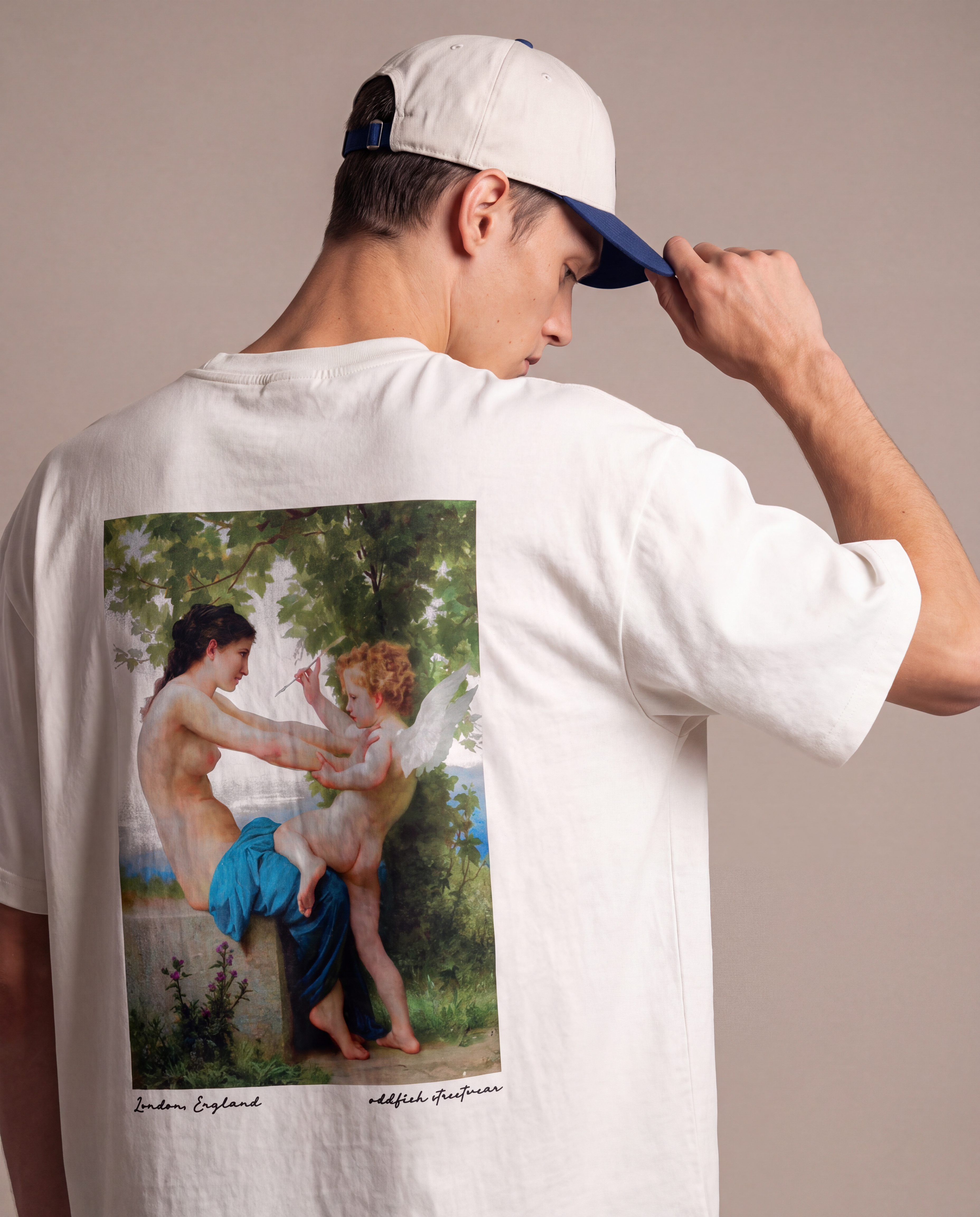A man wearing a white t-shirt with a large classical painting print on the back, shown from behind. He is also wearing a white baseball cap with a blue visor, set against a plain beige studio background.