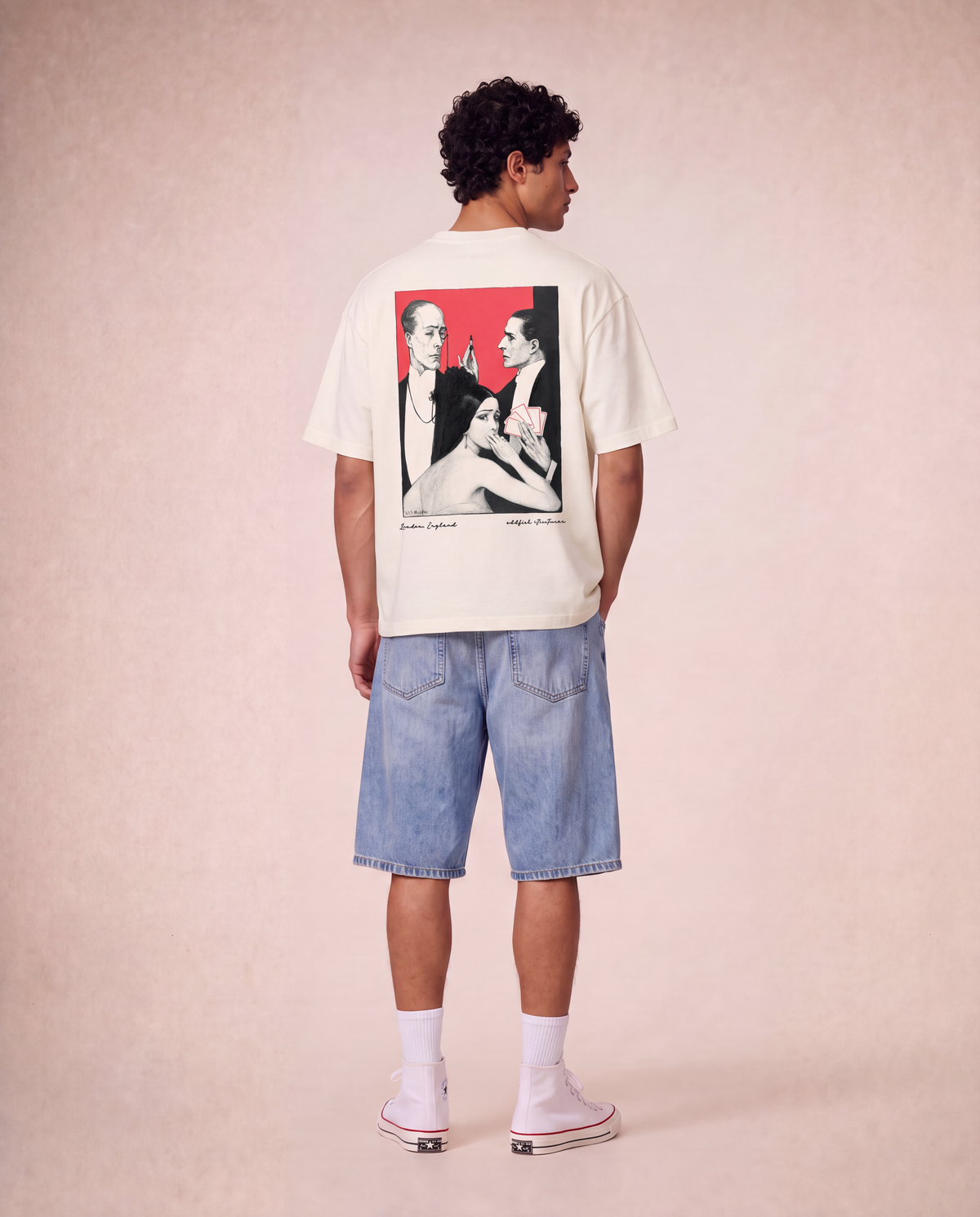 A model shown from the back wearing a cream-colored t-shirt with a large graphic print on the back, paired with light-wash denim shorts, white socks, and pink sneakers, set against a solid beige studio backdrop.
