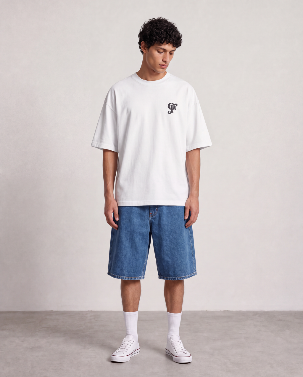 A man stands in a studio wearing a white oversized T-shirt with a small black chest logo, blue denim shorts, white socks, and white sneakers against a light neutral wall and gray floor.