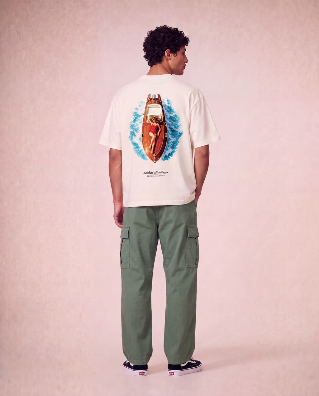 A man is shown from the back wearing a cream-colored t-shirt with a large graphic of a red speedboat on blue waves, paired with olive green cargo pants and black sneakers, set against a textured pink studio background.