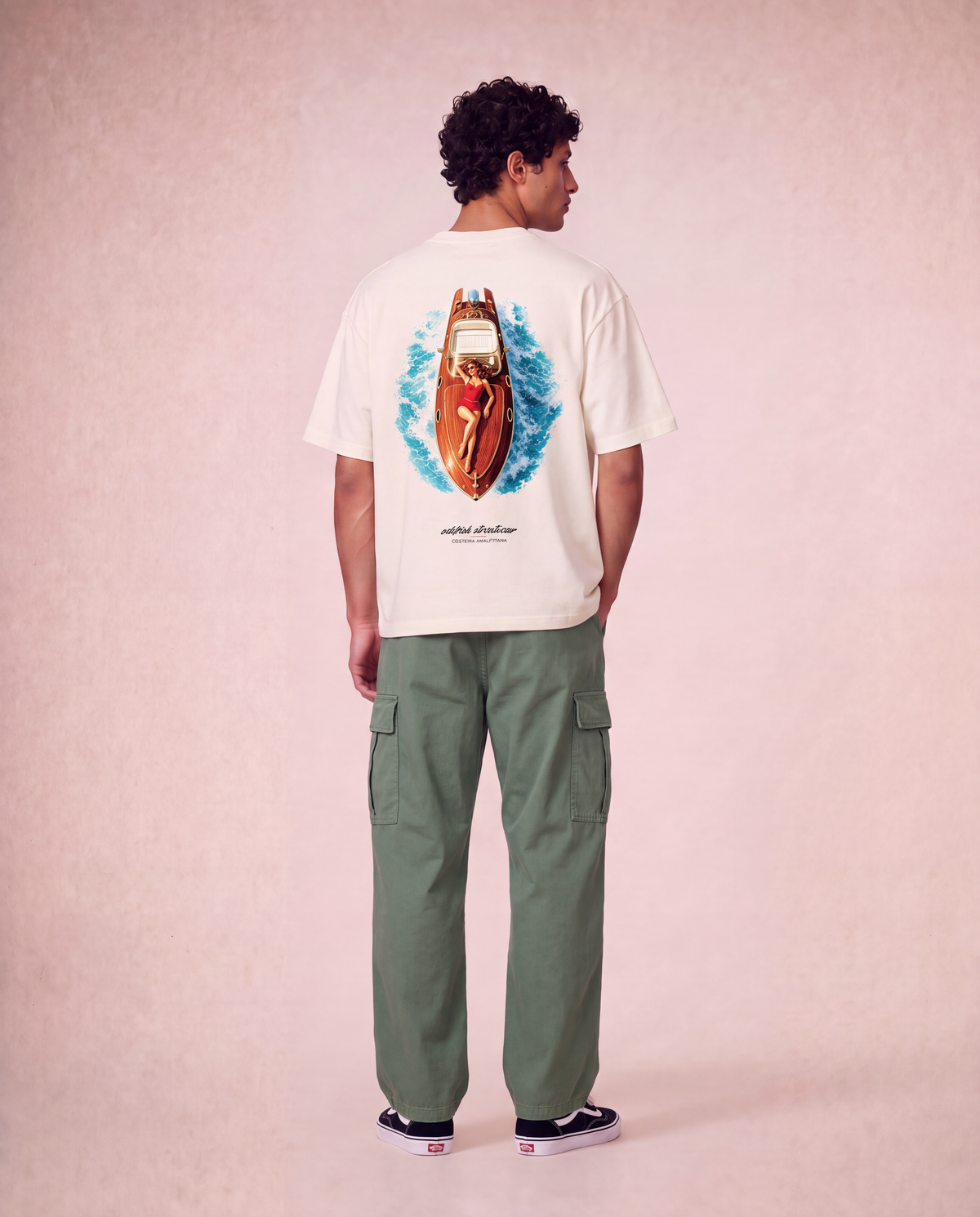 A man is shown from the back wearing a cream-colored t-shirt with a large graphic of a red speedboat on blue waves, paired with olive green cargo pants and black sneakers, set against a textured pink studio background.