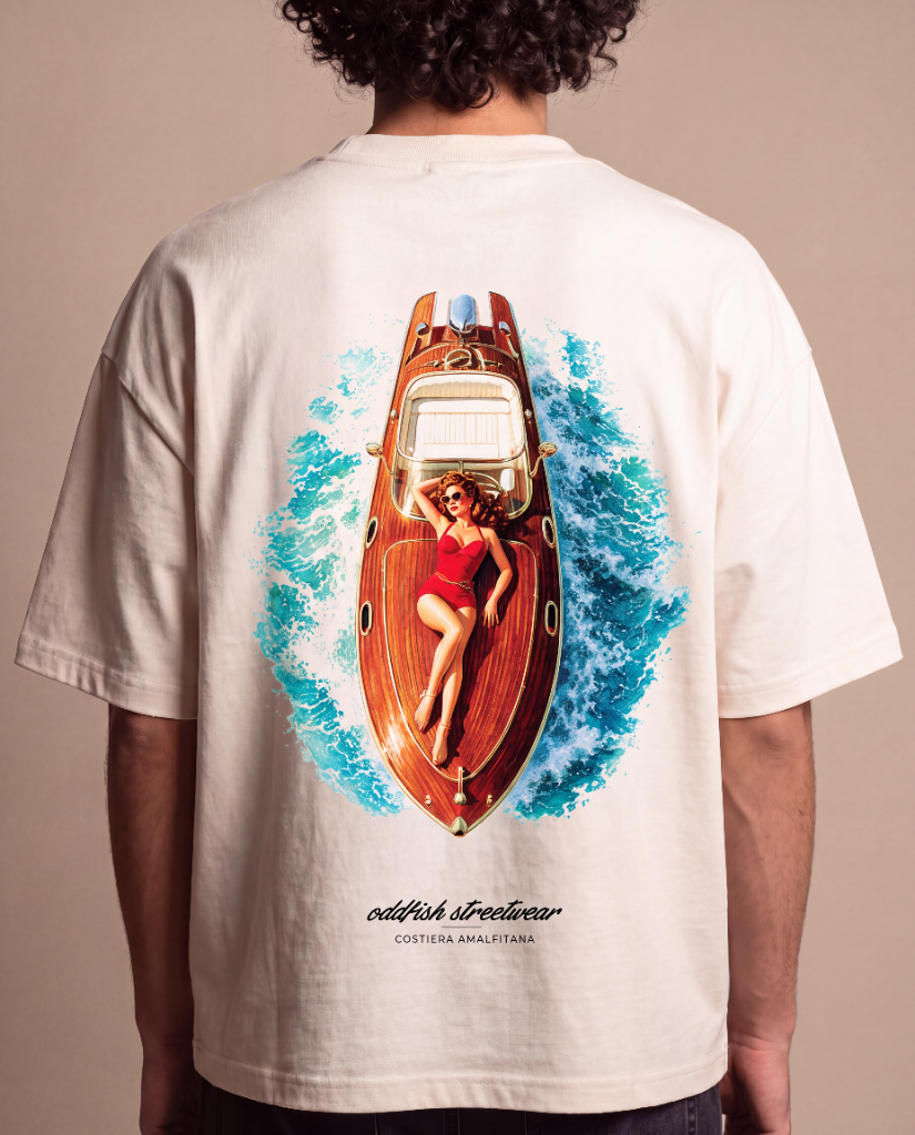 A person shown from the back wearing a cream-colored t-shirt with a large graphic print of a woman in a red swimsuit on a wooden yacht surrounded by blue waves. The text 'oddfish streetwear' and 'COSTESA ANALITICA' is printed below the graphic.