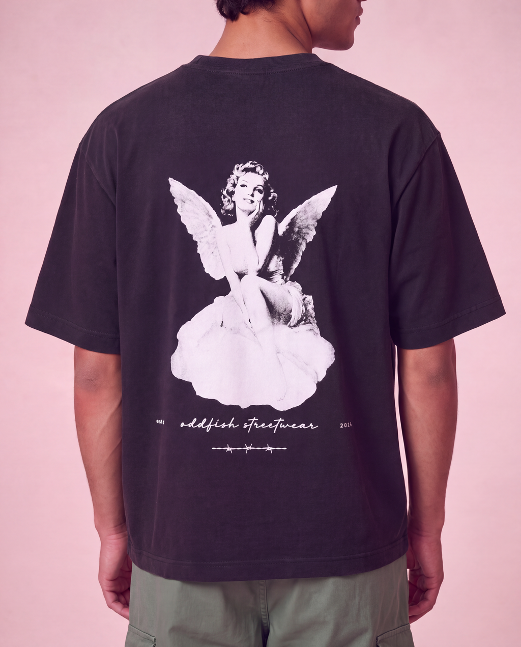 A person shown from the back wearing a dark charcoal t-shirt with a large white graphic of an angel on the back, paired with olive green pants, set against a solid pink background.