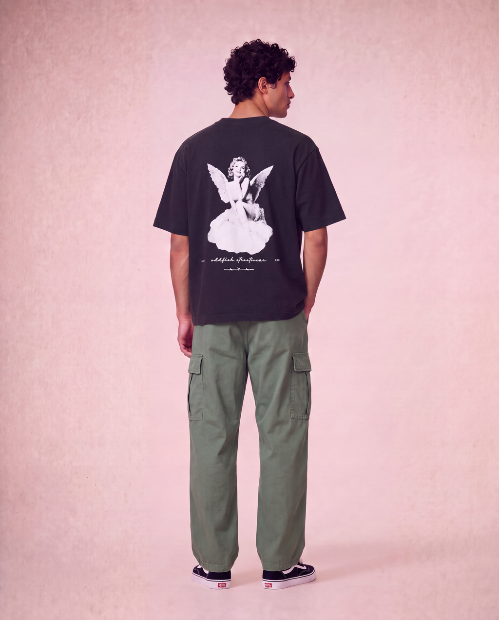 A man stands with his back to the camera wearing a black graphic t-shirt with a white angel illustration and olive green cargo pants, set against a textured pink studio backdrop.