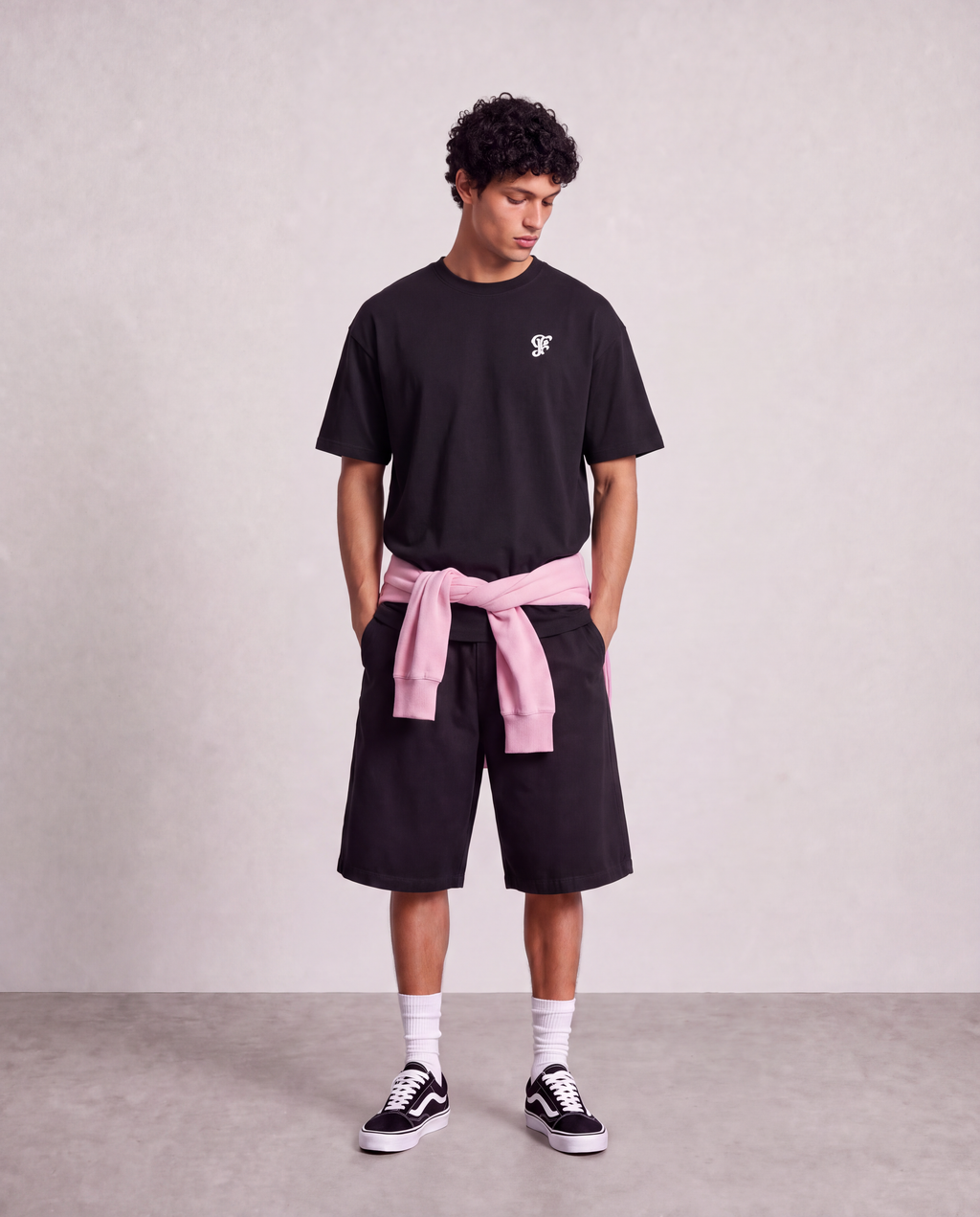 A man stands against a textured light gray wall wearing a black short-sleeve t-shirt with a small white chest logo, black shorts, white socks, and black-and-white sneakers. A light pink sweatshirt is tied around his waist like a belt.