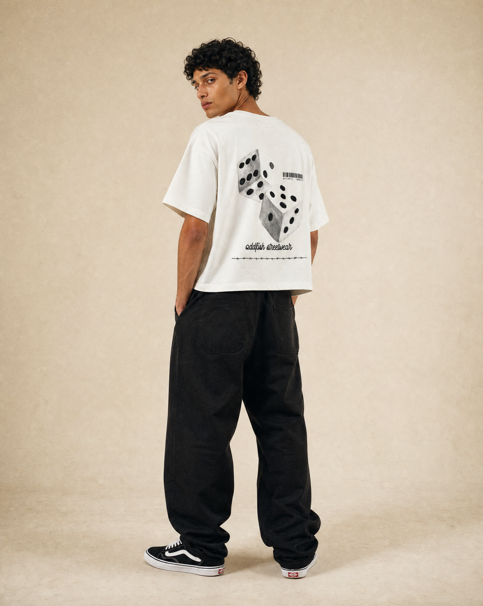 A man stands with his back to the camera wearing a white t-shirt with a large black graphic print on the back, black baggy pants, and black and white sneakers, set against a plain beige studio background.